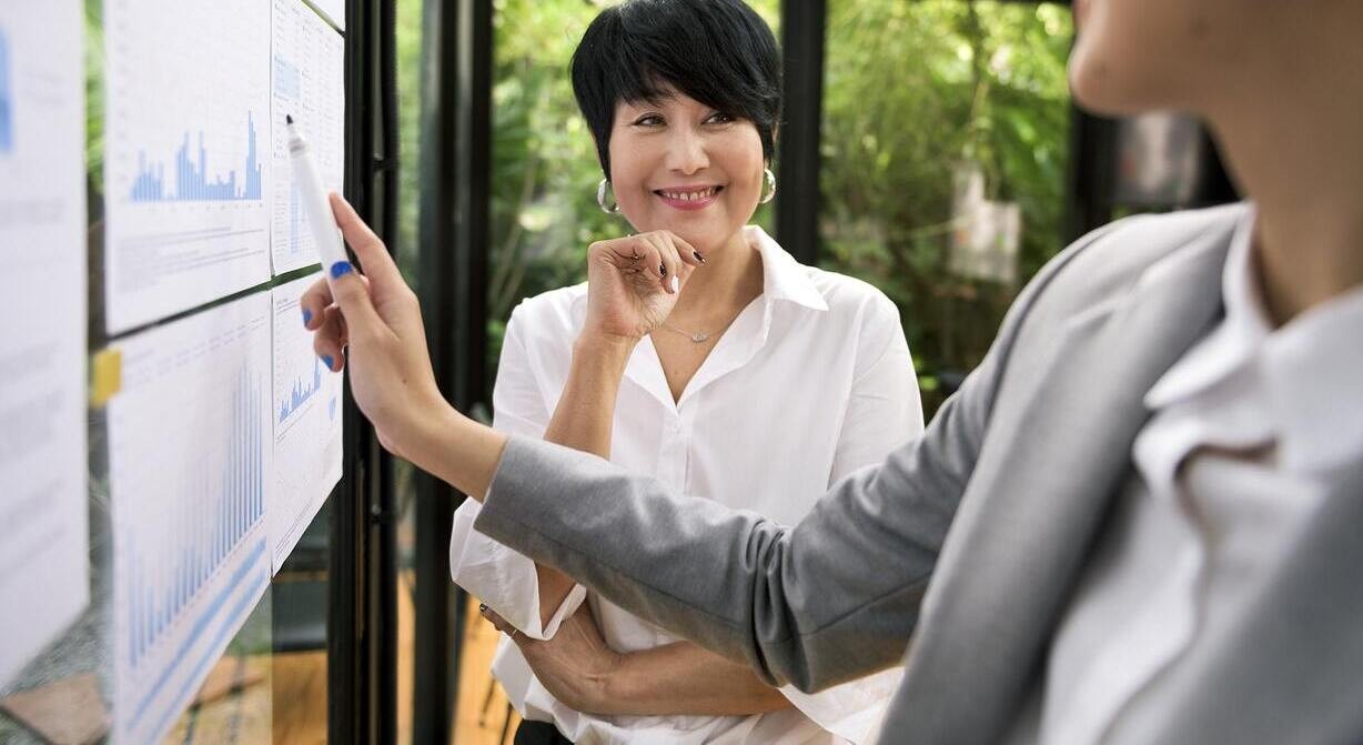 woman smiling while whatching at another woman point a chart on a board